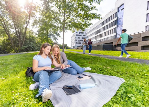 Am Gesundheitscampus startet zum Wintersemester 2026/27 der neue Bachelorstudiengang Gesundheitspsychologie. Zwei Studentinnen sitzen auf der Wiese am Gesundheitscampus und schauen in einen Ordner mit Unterlagen hinein.
