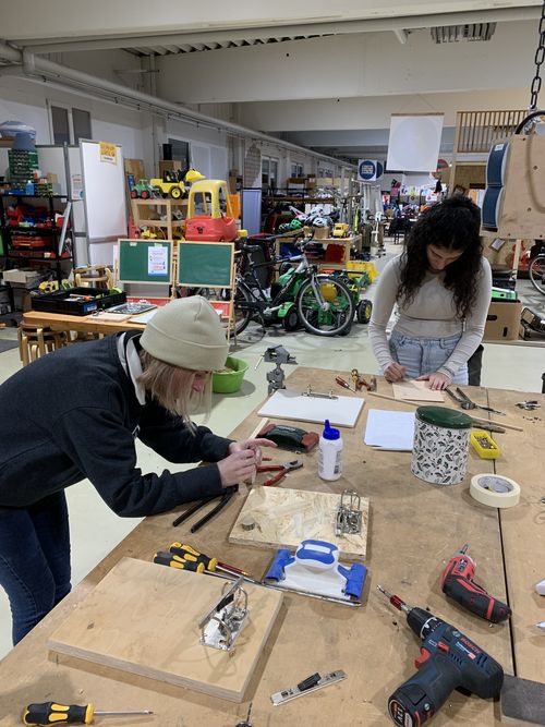 Foto vom Upcycling Workshop. 2 Teilnehmerinnen stehen um einen Werktisch und arbeiten an ihren Produkten