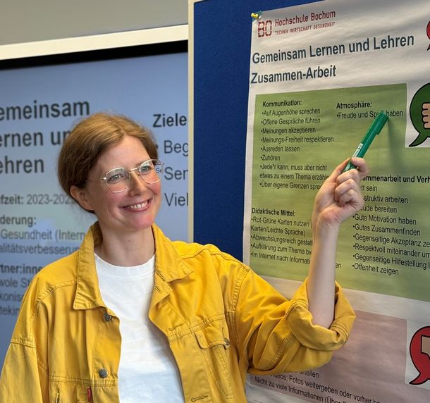 Julia Brüggemann zeigt auf ein Poster zum Projekt Gemeinsam lernen und lehren, das die Zusammenarbeit thematisiert.