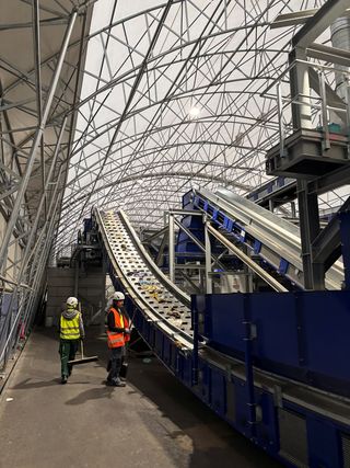 Zwei Personen mit Helm und Sicherheitsweste vor einem Förderband in einer großen Halle.
