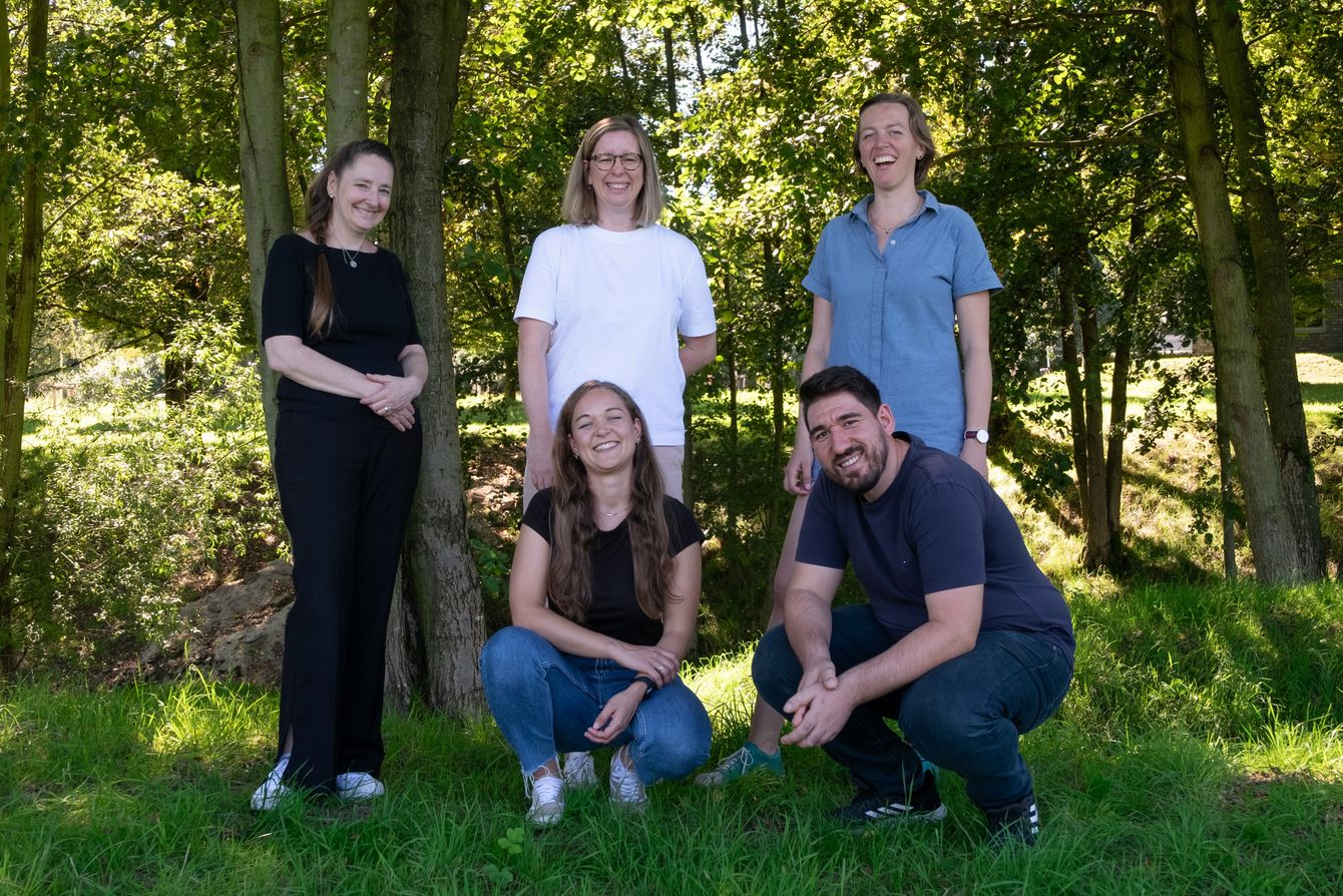 "Team photo of the founding team. Lenka, Lisa, and Judith are standing. Katharina and Anil are kneeling in front. In the background, there are trees and bushes