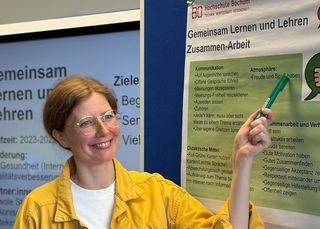 Julia Brüggemann zeigt auf ein Poster zum Projekt Gemeinsam lernen und lehren, das die Zusammenarbeit thematisiert.