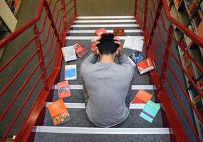 Rückenansicht eines Mannes, der auf einer Treppe sitzt und den Kopf in den Händen vergräbt, drumherum Bücher auf den Stufen