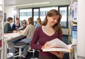 Studierende in der Hochschulbilbliothek. Eine Studierende im Vordergrund mit einem Buch zur Recherche in der Hand. Im Hintergrund ist eine Gruppe von fünf Studierenden, die an einem Stehtisch gemeinsam an einer Aufgabe arbeiten und eine Plakatpräsentation erstellen.
