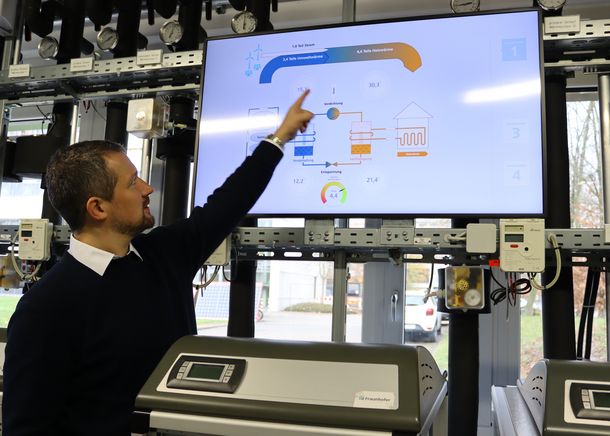 Prof. Dr. Rath zeigt auf einen Bildschirm, auf dem ein Beispiel für die Sektorenkopplung zu sehen ist: Der gewonnene Strom aus erneuerbaren Energien wie Wind und Sonne wird von Wärmepumpen zum Heizen genutzt.
