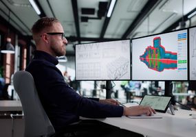 Ein Ingenieur arbeitet am Schreibtisch vor drei Monitoren mit 3D-CAD-Modellen und technischen Zeichnungen.