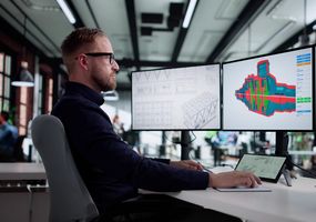 Ein Ingenieur arbeitet am Schreibtisch vor drei Monitoren mit 3D-CAD-Modellen und technischen Zeichnungen.