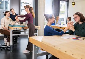 Studierende in der Ergotherapie-Werkstatt. Drei Tische mit je zwei Studierenden im Übungsseting. Eine Dozierende beobachtet die Situation.