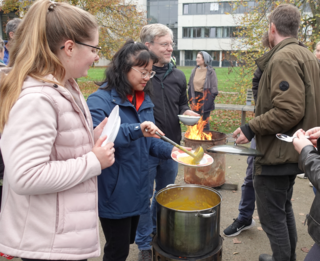 Die Abbildung zeigt Studierende und Mitarbeitende der Hochschule, die rund um den Pyrolyseofen verteilt stehen, auf dem die Kürbissuppe erhitzt wird.