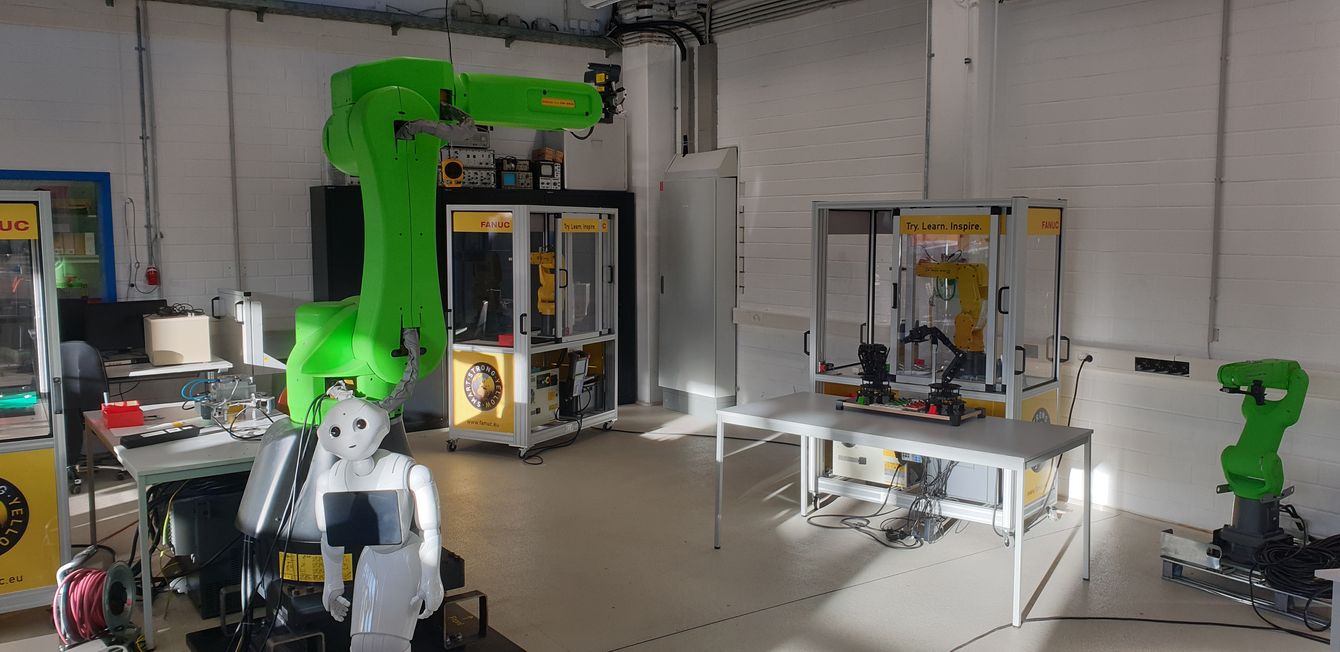 Ein grüner Roboter-Arm stehend im Labor umgeben mit Automaten und einem menschlichen Roboter im Vordergrund