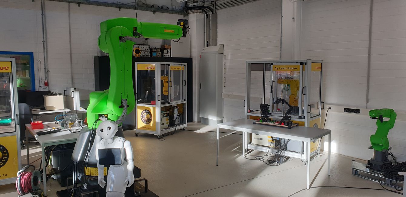 Ein grüner Roboter-Arm stehend im Labor umgeben mit Automaten und einem menschlichen Roboter im Vordergrund Ein grüner Roboter-Arm stehend im Labor umgeben mit Automaten und einem menschlichen Roboter im Vordergrund